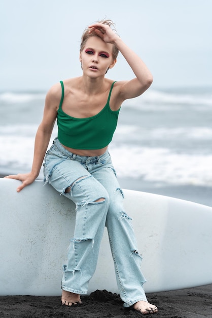 Foto mulher vestindo top e jeans azuis sentada em uma prancha de surf deitada na areia no fundo das ondas do mar