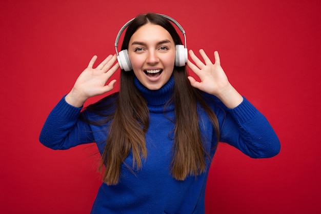 mulher vestindo um suéter azul isolado sobre a parede de fundo vermelho usando fones de ouvido bluetooth brancos, ouvindo música legal e se divertindo olhando para a câmera.