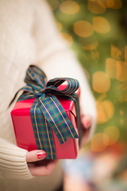 Mulher vestindo um suéter e luvas vermelhas sazonais contra um fundo abstrato verde e dourado, segurando um presente de Natal lindamente embrulhado com profundidade de campo estreita