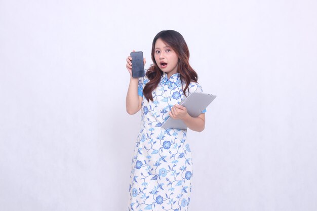 mulher vestindo vestido chinês olhando para a câmera chocado segurando tablet e olhando para o ecrã do telefone celular