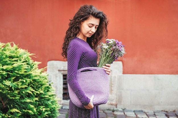 Mulher vestindo vestido de bolinhas roxas segurando saco de malha com flores posando na rua da cidade