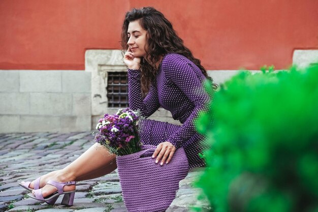 Mulher vestindo vestido de bolinhas roxas segurando saco de malha com flores posando na rua da cidade