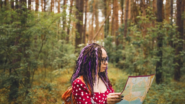 Mulher viajando com dreadlocks com mapa na floresta Viajante feminina com mochila na floresta lendo mapa em tempo nublado