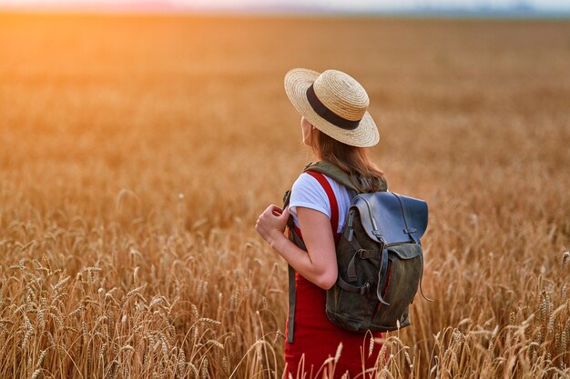 Mulher viajante com mochila sozinha no campo de trigo seco amarelo dourado com espigas desfrutando de um lindo momento de liberdade no verão ao pôr do sol