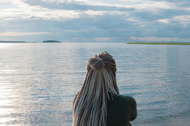 mulher viajante com penteado de tranças sentada em uma praia de areia de um vasto lago