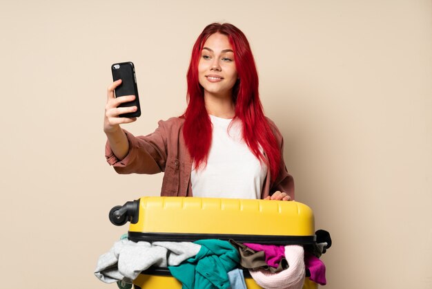 Mulher viajante com uma mala cheia de roupas isoladas na parede bege, fazendo um selfie
