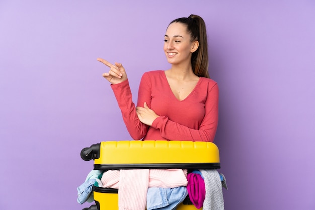 Mulher viajante com uma mala cheia de roupas sobre parede roxa isolada, apontando o dedo para o lado