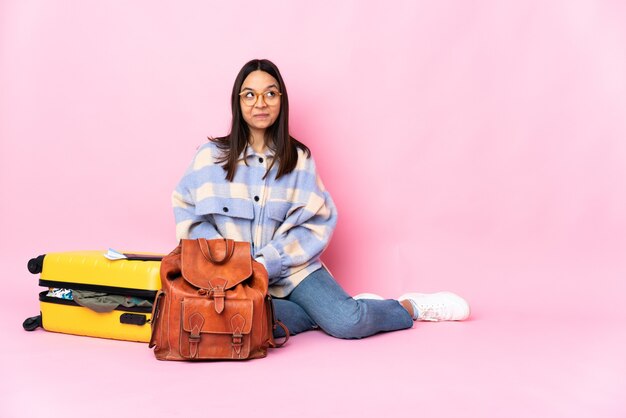 Mulher viajante com uma mala sentada no chão, tendo dúvidas enquanto olha para cima