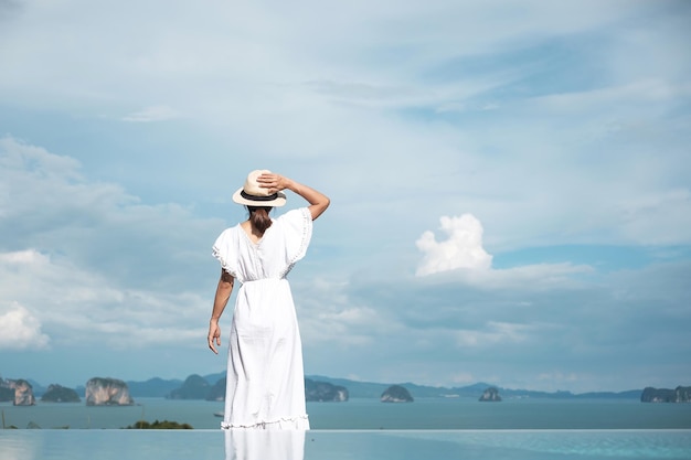 Mulher viajante feliz de vestido branco desfruta de uma bela vista sozinha Turista com chapéu em pé e relaxando sobre o conceito de verão e férias de viagem de piscina