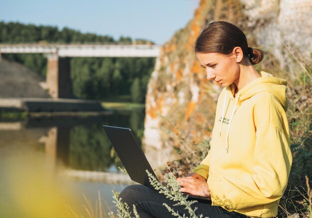 Mulher viajante freelancer com capuz amarelo com laptop aberto no fundo da ponte e do rio