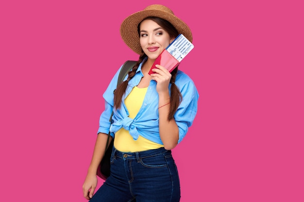 Mulher viajante sorridente com chapéu de palha e mochila segurando o passaporte com passagem de avião sobre fundo rosa isolado