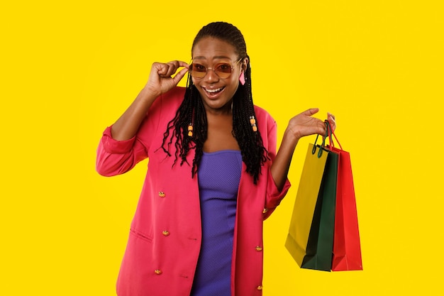 Mulher viciada em compras africana usando óculos escuros segurando sacolas de compras fundo amarelo