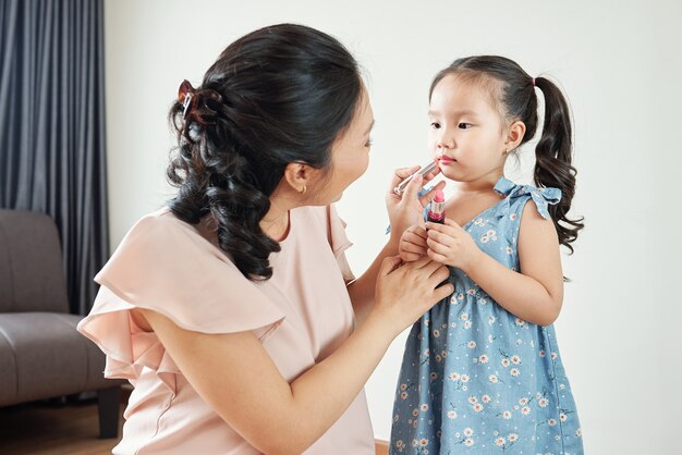 Mulher vietnamita sorridente aplicando bálsamo labial hidratante rosa claro nos lábios de sua filha