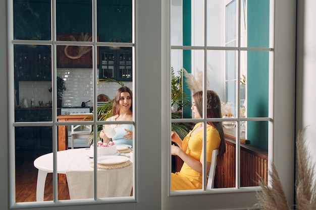 Mulheres amigas felizes em casa sentadas e sorrindo com bolo de aniversário branco atrás da porta de vidro.