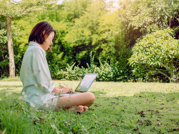 Mulheres asiáticas bonitas que vestem a camisa e o short brancos com o laptop que senta-se na grama verde.