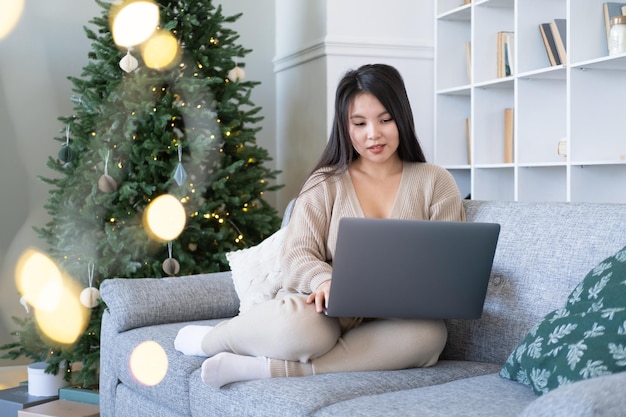 mulheres asiáticas jovens atraentes em roupas aconchegantes usando laptop no sofá em casa, tempo de natal