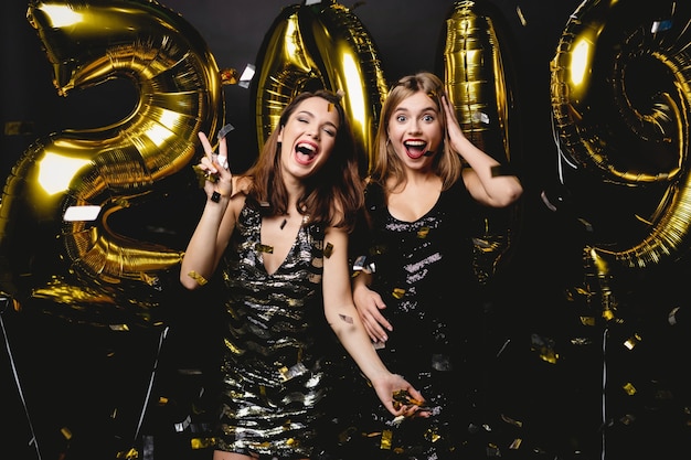 Mulheres bonitas comemorando o ano novo. Garotas lindas e felizes em vestidos de festa sensuais elegantes segurando balões de ouro de 2019, se divertindo na festa de ano novo. Celebração do feriado. Imagem de alta qualidade