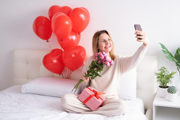 Mulheres bonitas seguram buquê de rosas cor de rosa tira selfie no telefone na cama comemorando o dia dos namorados