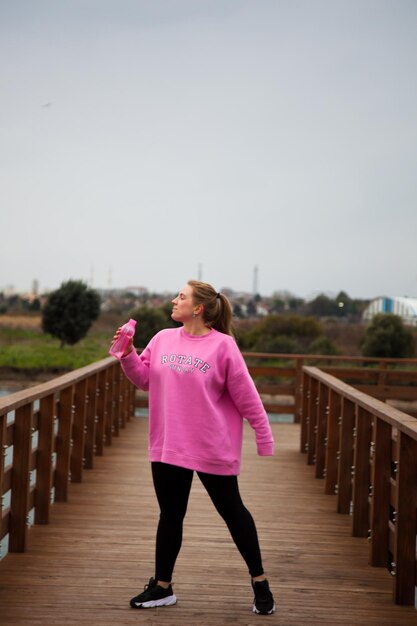 Foto mulheres com capuz rosa fazendo exercícios ao ar livre no cais no outono. mulher bebendo água após exe