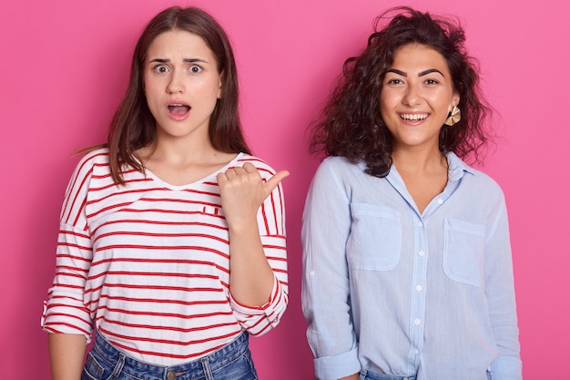 mulheres de cabelos escuros, adorável senhora com a boca aberta aponta o polegar para sua amiga sorridente, as fêmeas usam roupas casuais