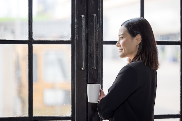 Mulheres de negócios asiáticas segurando uma xícara de café e em pé perto da janela do escritório no intervalo