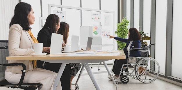 Mulheres de negócios asiáticas sentadas na mesa com laptops na frente, ouvindo a mulher de negócios com deficiência sentada na cadeira de rodas, apresentando tabelas e gráficos no quadro no escritório.