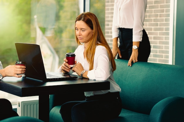 Mulheres de negócios em trajes formais elegantes estão discutindo assuntos, usando um laptop, bebendo café e sorrindo enquanto trabalham juntos no café. Reunião de negócios em um novo e moderno ambiente de trabalho em tijolos à vista