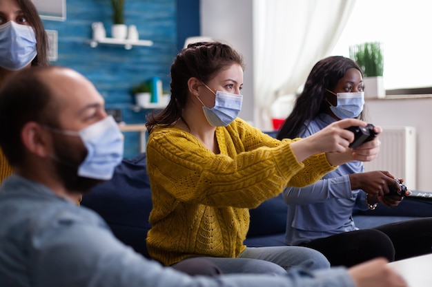 Mulheres de raça mista com máscara de proteção jogando videogame segurando o controle sem fio desfrutando de uma nova festa normal relaxante com amigos. diversas pessoas mantendo o distanciamento social contra a pandemia global