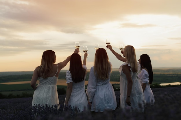 Mulheres de vestido branco erguem seus copos para brindar ao pôr do sol