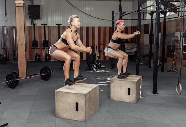 Mulheres desportivas fazendo saltos de caixa juntos no ginásio. Aula de treinamento pulando em caixas de madeira no clube de saúde cross fit.