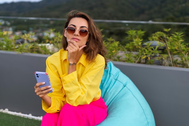 Mulheres elegantes da moda em calças largas rosa brilhante e camisa amarela segurando bolsa óculos de sol da moda posando no terraço da cobertura vista tropical ao ar livre segurando o telefone móvel