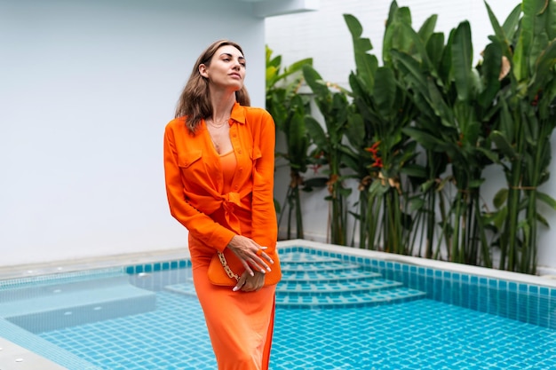 Mulheres elegantes da moda em vestido laranja brilhante e camisa segurando bolsa de moda posando em villa tropical de luxo à beira da piscina luz natural do dia ao ar livre