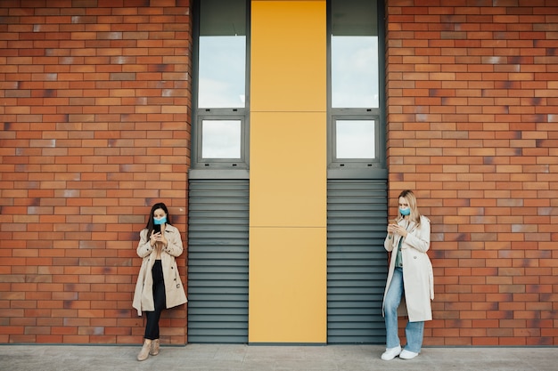 Mulheres elegantes usando máscaras estão usando smartphones perto de uma parede de tijolos.