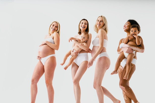 Mulheres grávidas felizes com barriga grande e lindas mães com bebês em estúdio - grupo multirracial de mulheres grávidas e mães com crianças vestindo roupas íntimas - gravidez, maternidade, pessoas, postura corporal