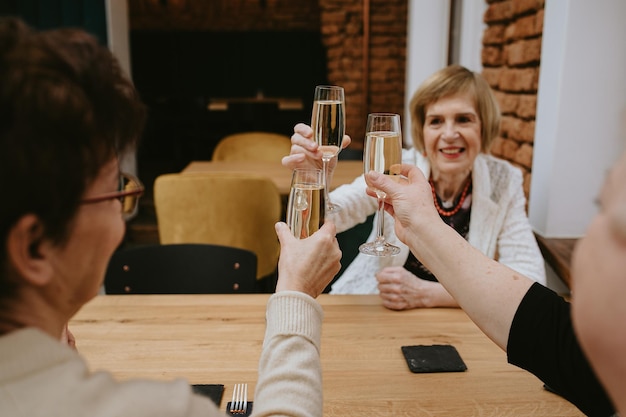 Mulheres idosas de cabelos escuros e louros sorridentes com maquiagem vestida com jaquetas pretas e brancas brindando com taças de vinho ou champanhe no café comemorando sua reunião com amigos