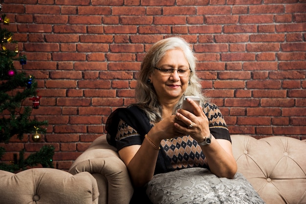 Mulheres indianas asiáticas sêniors ouvindo música usando fones de ouvido enquanto seguram um computador tablet ou usam ou falam no telefone e estão sentadas no sofá ou sofá em casa