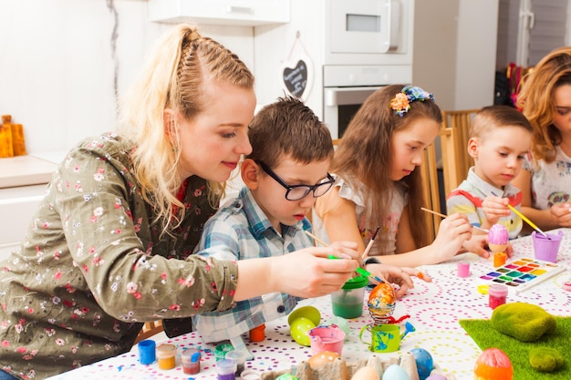 Mulheres jovens ajudando crianças criando uma decoração de primavera para a Páscoa. Mãe e filho segurando pincéis pintando ovo colorido