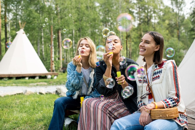 Mulheres jovens e multiétnicas positivas sentadas em uma barraca na floresta e soprando bolhas de sabão