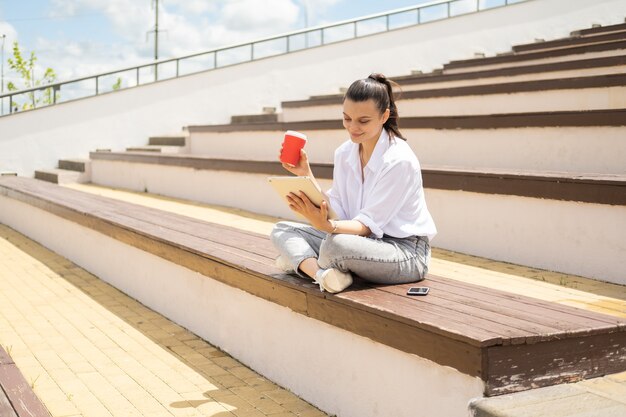 Mulheres jovens felizes com um tablet segurando um copo de papel de café, aproveitando o dia de sol sentado no anfiteatro