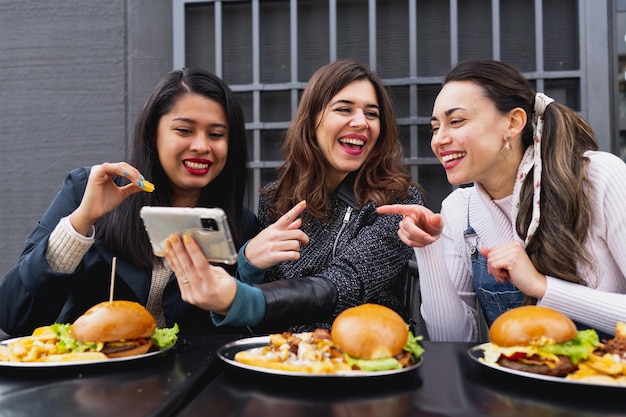 Mulheres jovens felizes sentadas à mesa comendo hambúrgueres e batatas fritas Mulheres rindo
