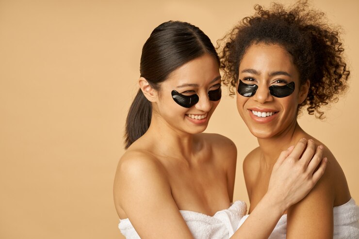 Mulheres jovens sorridentes, enroladas em uma toalha, posando com hidrogel preto aplicado sob o tapa-olho