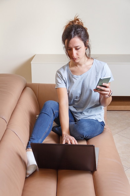 Mulheres jovens trabalhando com laptop e usando o celular, sentadas no sofá