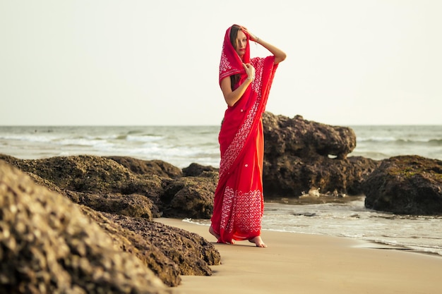 Mulheres jovens vestindo um saree vermelho na praia goa india.girl em sari indiano tradicional na costa de uma ilha paradisíaca entre as rochas e areia, apreciando a liberdade e o pôr do sol.