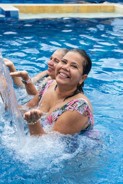 Mulheres maduras sorridentes com cabelo molhado e biquíni sob o jato de água na piscina
