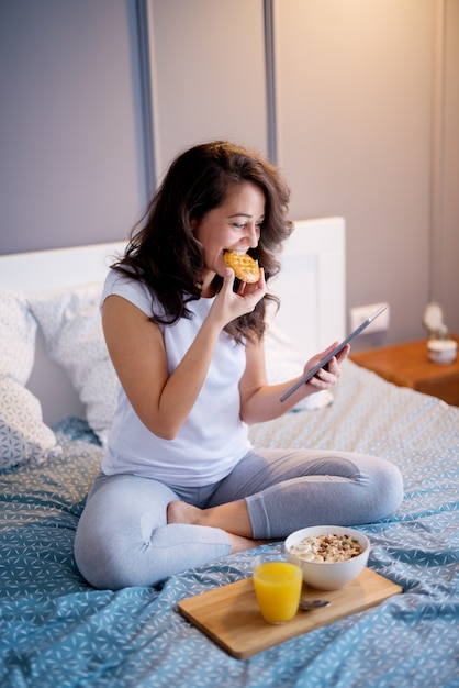 Mulheres meio envelhecidas consideravelmente satisfeitas que sentam-se na cama e que olham em uma tabela antes de dormir ao comer um biscoito.