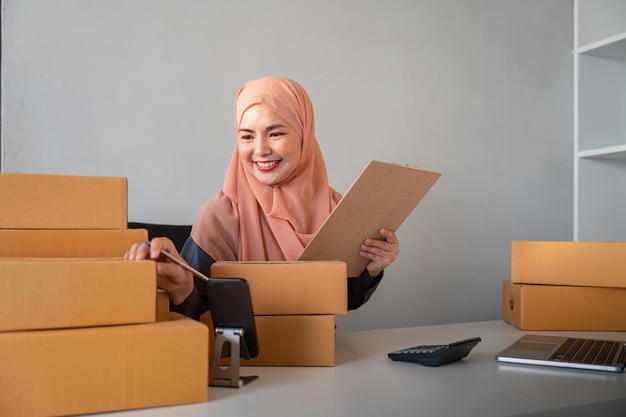 Mulheres muçulmanas vendendo online em casa com caixa Vendo online com caixa para aceitar encomenda do cliente ideia de negócio de PME Entrega de encomendas mulher muçulmana trabalhando com smartphone e laptop em casa.