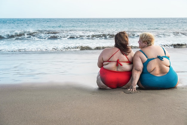 Mulheres plus size sentadas na praia se divertindo durante as férias de verão - Retrovisor de uma mulher cheia de curvas rindo juntas - Sobrepeso e conceito de corpo diversificado