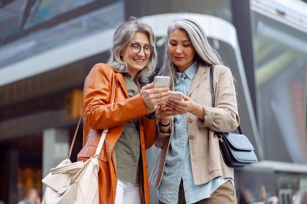 Mulheres positivas de cabelos grisalhos olham para telefones celulares nas ruas da cidade moderna