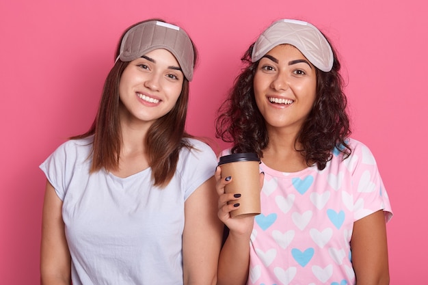 Mulheres satisfeitas posando isoladas sobre fundo rosa, de pijama e máscaras de dormir, de bom humor, sorrindo para a câmera, expressando positivo, acordam depois da festa do pijama.