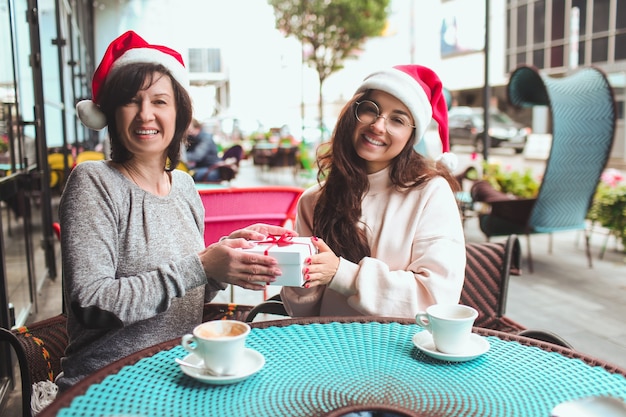 Mulheres sentadas juntas usando chapéus de Papai Noel e segurando um presente juntas